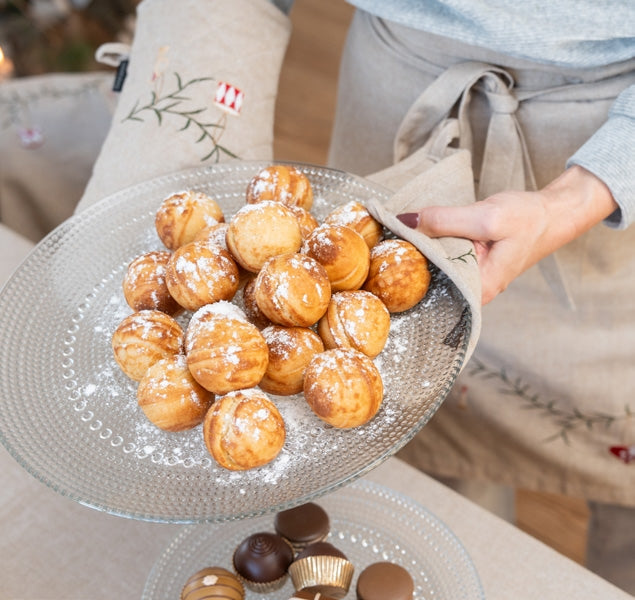 A person wearing a beige apron holds a glass plate filled with golden-brown fried pastries dusted with powdered sugar, with chocolate-covered pastries visible in a matching glass plate below on a table.