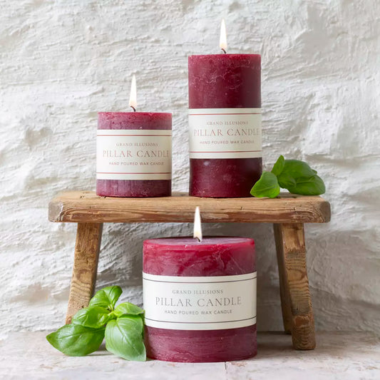 Three dark burgundy pillar candles with white paper labels reading 'GRAND BLESSINGS PILLAR CANDLE HAND POURED WAX CANDLE' are styled on and around a small rustic wooden stool against a white textured wall. Two candles are lit, and green basil leaves are placed near them.