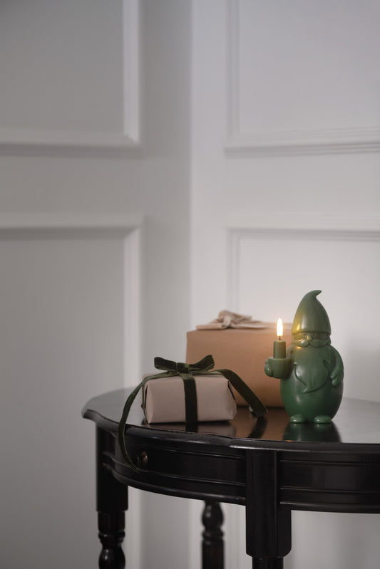 A green gnome-shaped candle holder with a lit candle sits on a dark wooden table next to a wrapped gift box and a brown box against a white paneled wall.