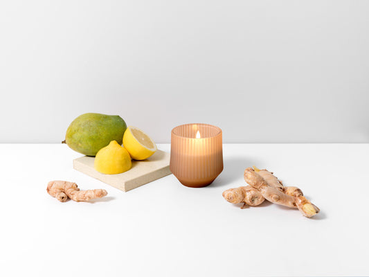 A lit candle in a ribbed amber glass jar sits on a white surface with ginger roots, a green mango, and yellow lemons arranged around it against a clean white background.