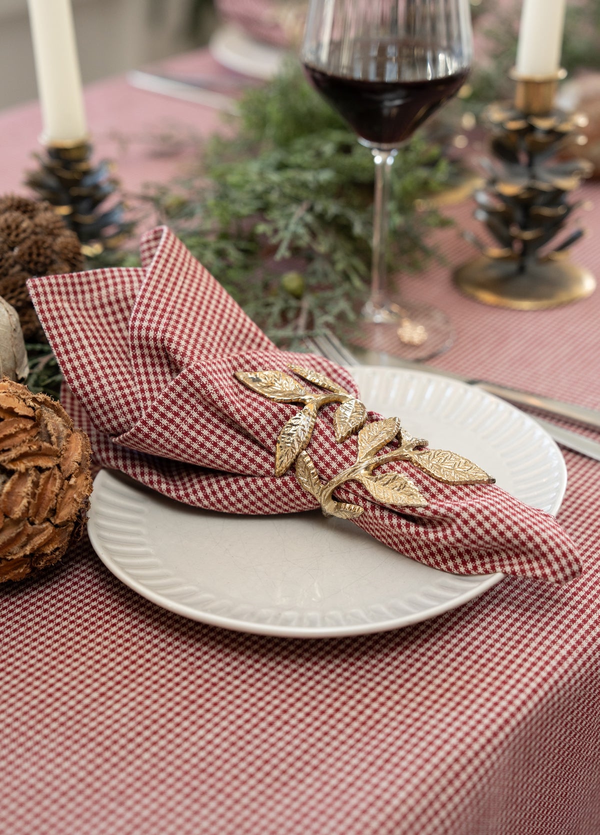 A close-up of a styled table setting featuring a red and white gingham check napkin held in place by a gold leaf-patterned napkin ring, set on a white plate over a matching checkered tablecloth.