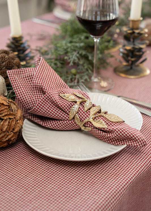 A close-up of a styled table setting featuring a red and white gingham check napkin held in place by a gold leaf-patterned napkin ring, set on a white plate over a matching checkered tablecloth.