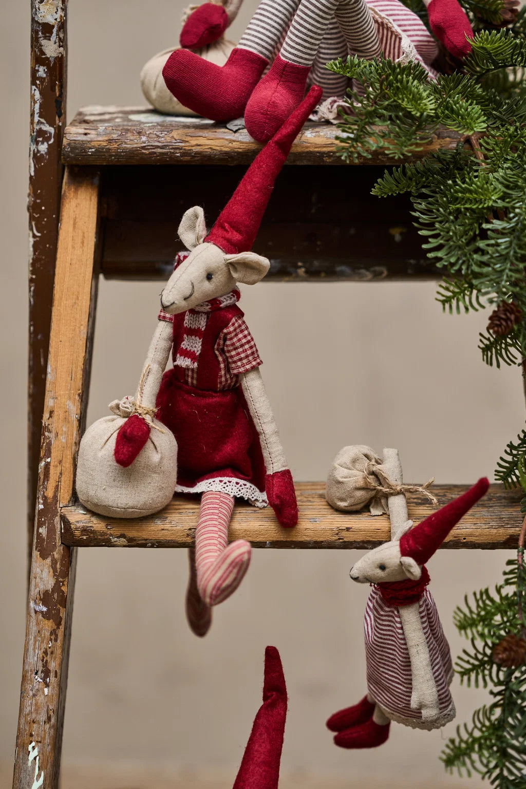 A rustic Christmas display featuring handmade felt mouse dolls with red pointed hats and dresses, arranged on a weathered wooden ladder alongside green pine branches against a neutral beige background.