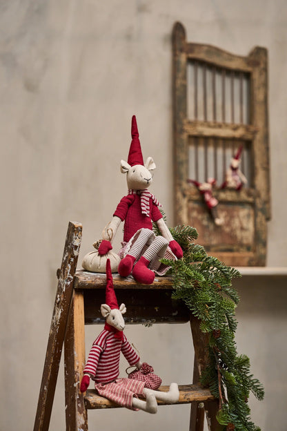 Two small fabric reindeer dolls dressed in red and white striped outfits and pointed red hats, posed on a rustic wooden ladder with green pine garland in a styled indoor setting.