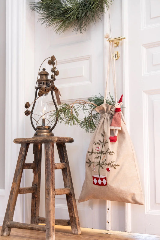 A rustic beige burlap Christmas stocking hangs from a brass hook on a white paneled door, decorated with a small embroidered Christmas tree and a red-and-white patterned pot. A wooden stool with a vintage lantern and pine branches sits beside it.