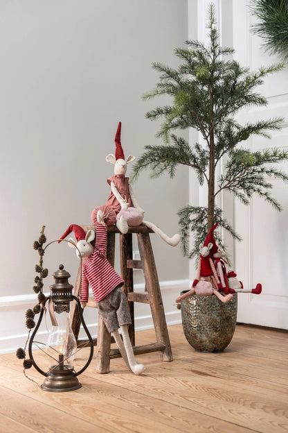 Three rustic Christmas mouse figurines dressed in red and white striped outfits and pointed hats, posed with a wooden stool, a vintage lantern, and a small potted tree on a light wood floor against a neutral gray wall.