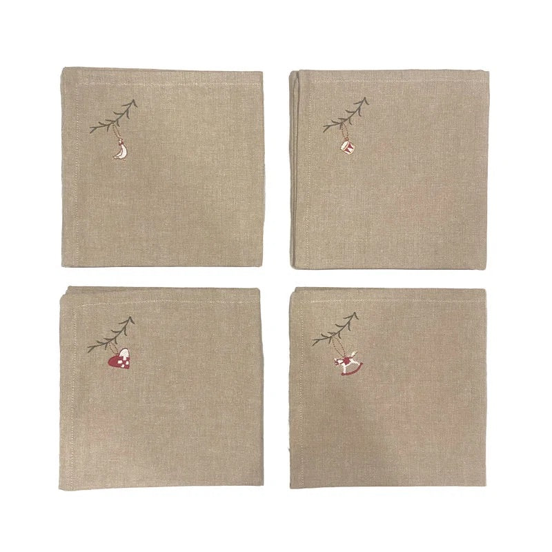 Four beige linen napkins arranged in a 2x2 grid on a white background. Each napkin features a small embroidered Christmas motif in the top-left corner, including a hanging ornament and a red-and-white stocking on a small branch.