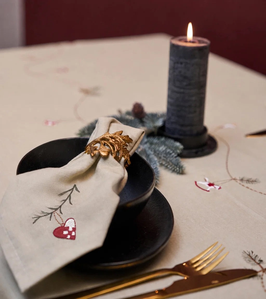 A styled table setting featuring a beige embroidered tablecloth with delicate floral and heart motifs, a black bowl and saucer, a cream napkin with a gold pinecone ring, gold flatware, and a lit dark gray candle.