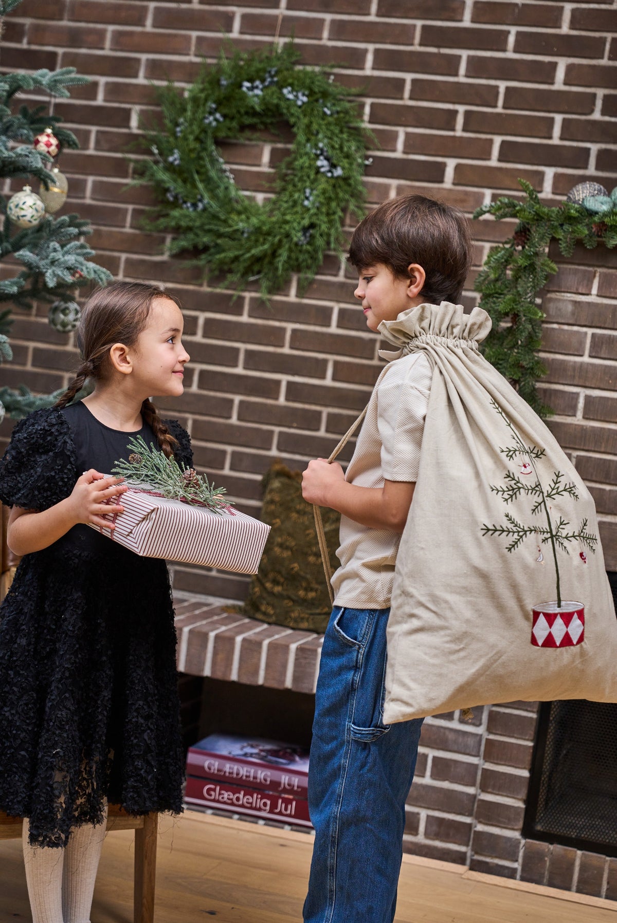 A young boy and girl standing in front of a brick fireplace. The boy wears a beige Christmas sack with a green tree design and holds a wrapped gift box, while the girl in a black lace dress holds a small green sprig.
