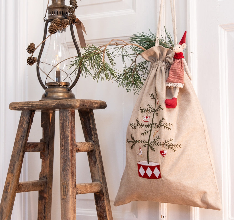 A beige fabric Christmas stocking bag with an embroidered pine tree and red-and-white ornaments, hanging on a white door next to a rustic wooden stool with a lantern and evergreen branch.