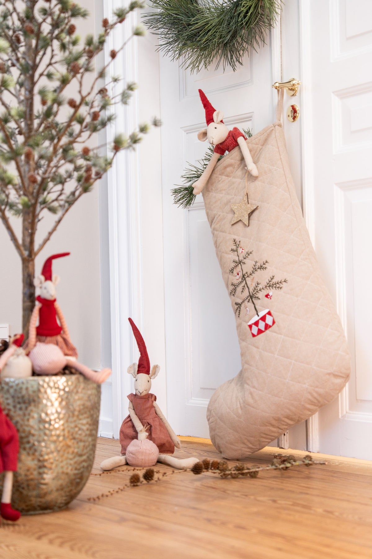 A beige quilted Christmas stocking with embroidered tree and star details hangs on a white door, styled with plush gnomes and holiday decor in a warm indoor setting.