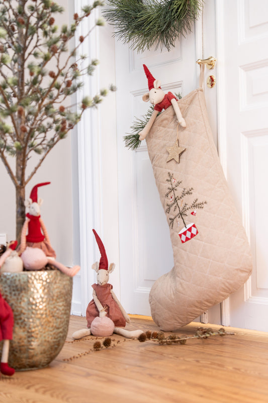A beige quilted Christmas stocking with embroidered tree and star details hangs on a white door, styled with plush gnomes and holiday decor in a warm indoor setting.