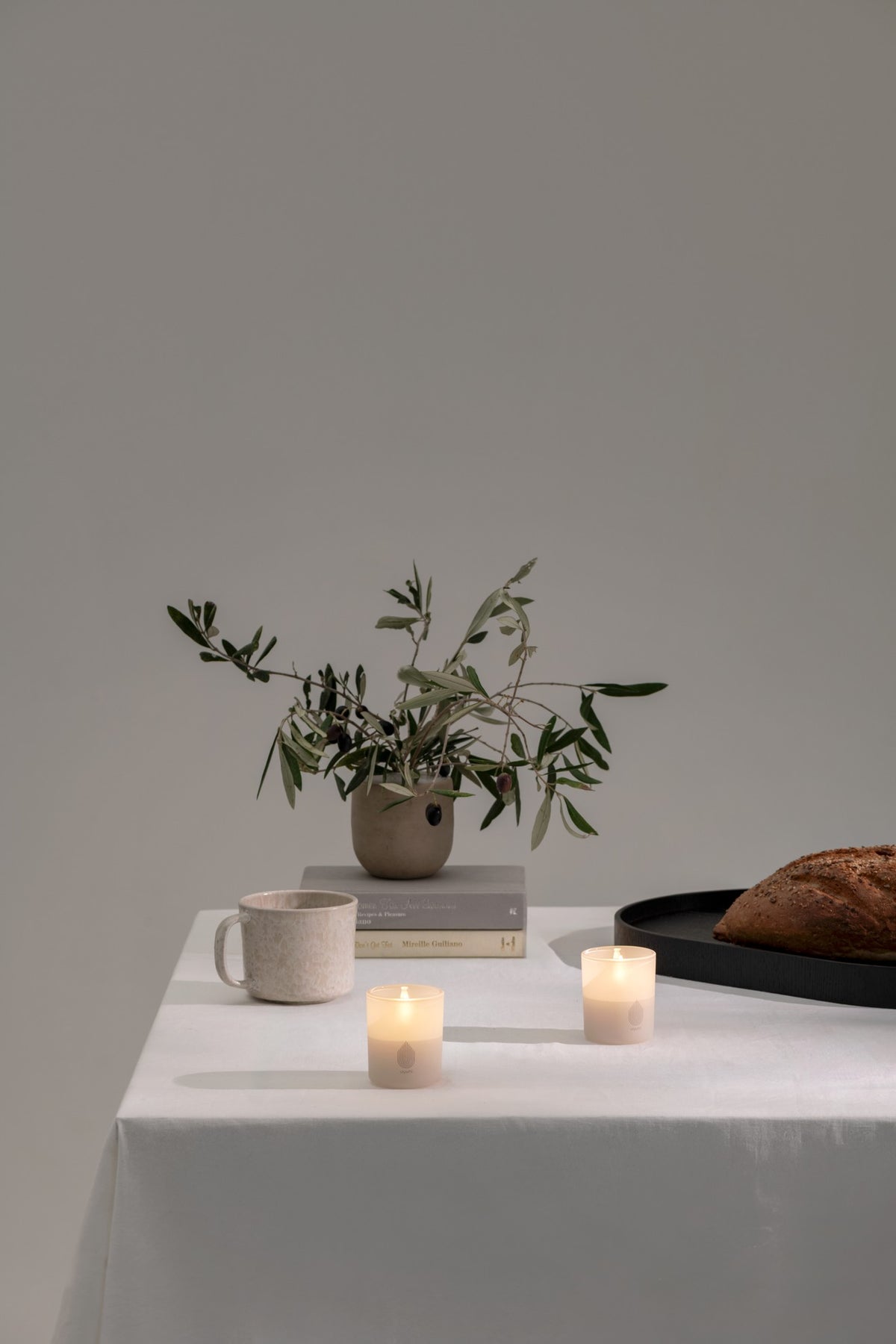 A styled tabletop scene with a white tablecloth, a small vase of greenery, stacked books, a ceramic mug, two lit candles, and part of a loaf of bread against a plain light gray wall.
