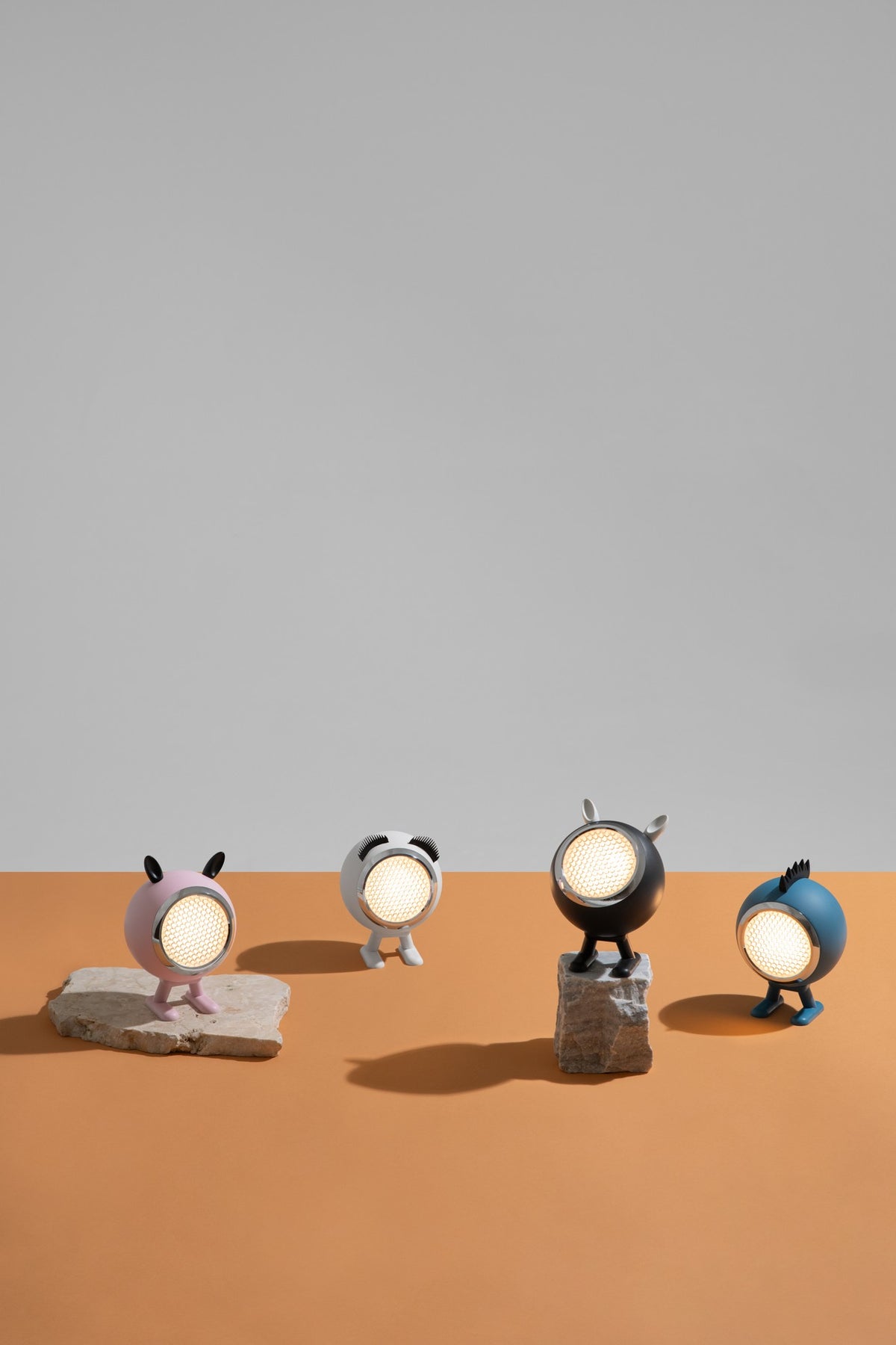 Four small character-shaped table lamps with round glowing faces stand on stone-like bases against a split gray and orange studio background.