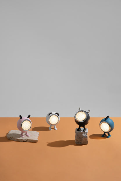 Four small character-shaped table lamps with round glowing faces stand on stone-like bases against a split gray and orange studio background.
