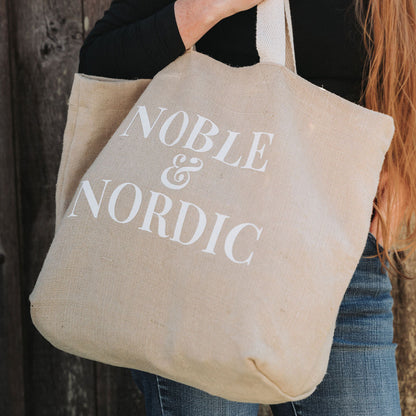 Person holding a beige tote bag with 'Noble & Nordic' text against a wooden background designed by Noble & Nordic