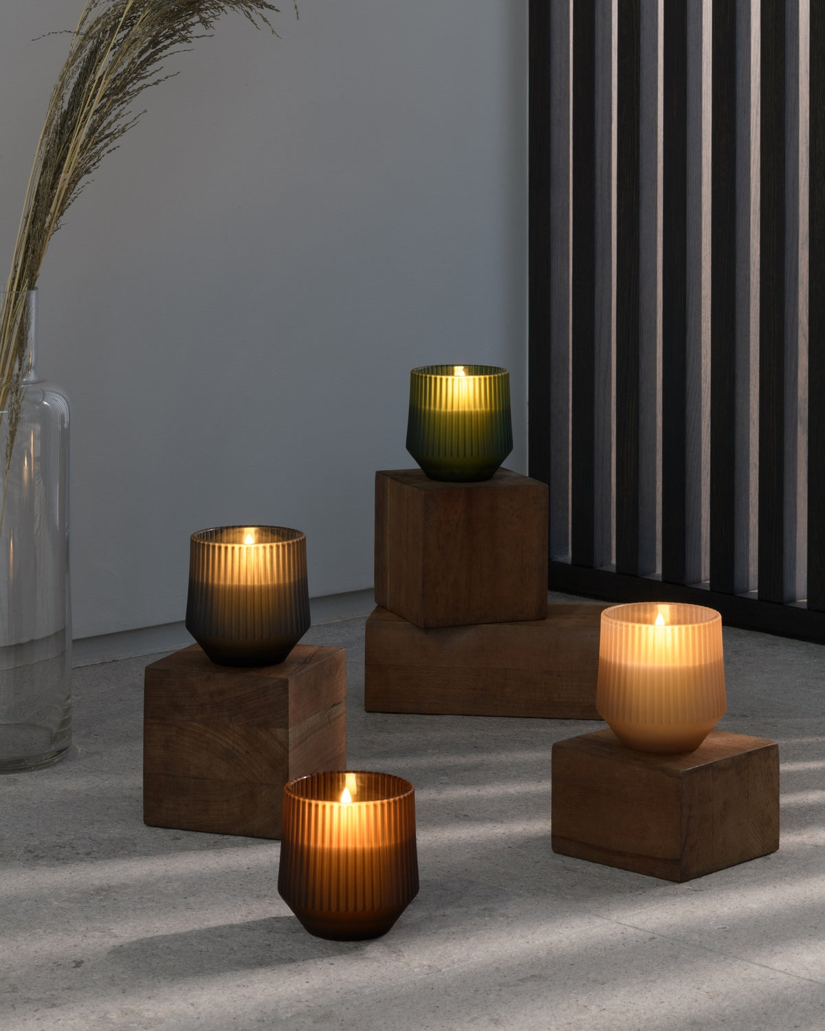 Four lit candle holders in different colors and textures arranged on wooden blocks in a styled interior setting with a gray wall, slatted screen, and dried grass in a glass vase.