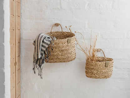 Two woven natural-fiber baskets hanging on a white brick wall, with a striped throw draped over the larger basket and dried stems in the smaller one.