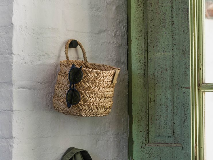 A woven straw basket bag with a top handle hangs on a white brick wall next to a distressed green wooden door. A pair of sunglasses is draped over the side of the bag.