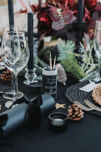 Decorative table setting with wine glasses, candles, and fir garland on a black tablecloth by Noble & Nordic.