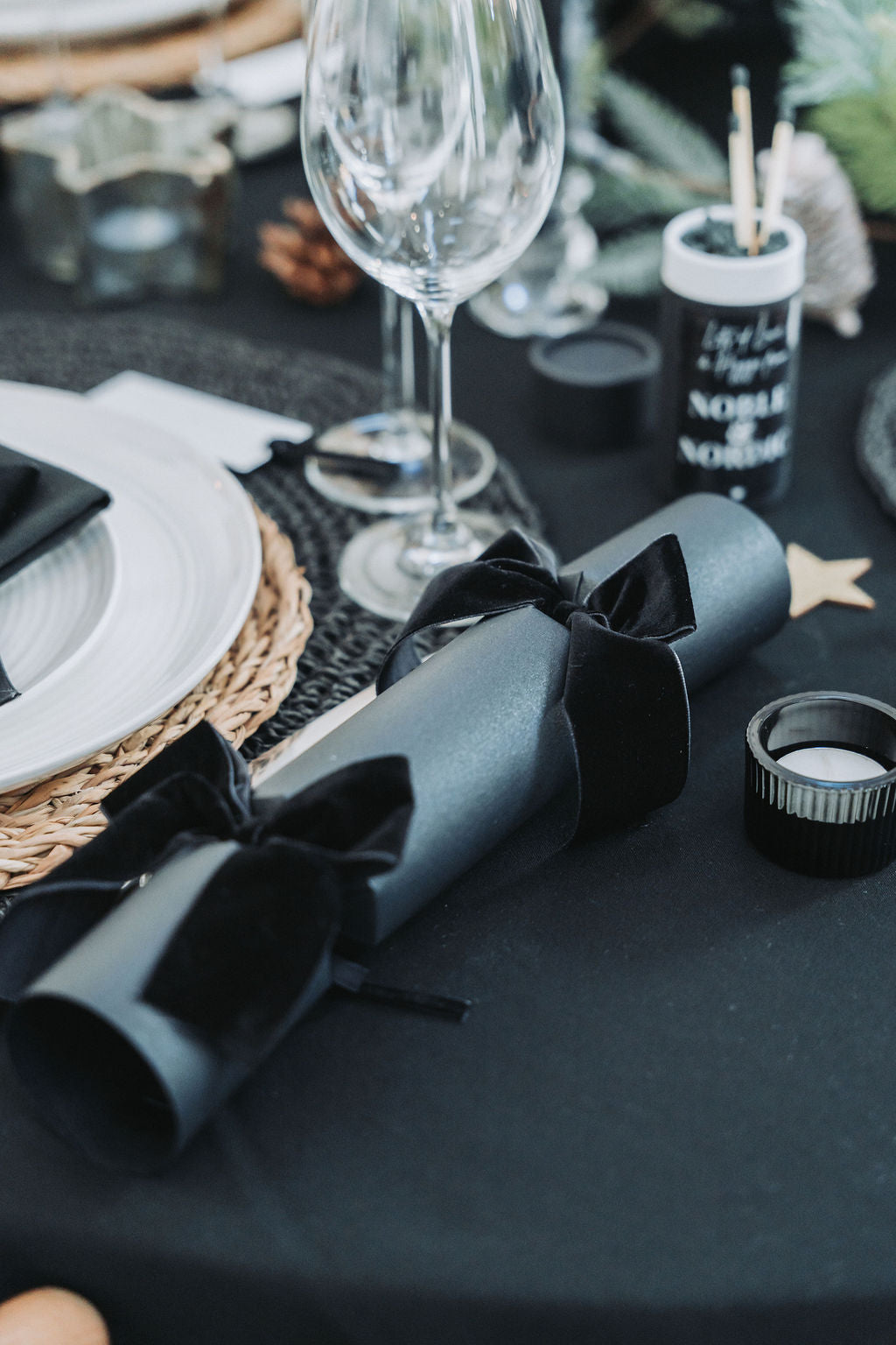 Decorative table setting with black napkins, glasses, and black candles on a black tablecloth with black Christmas crackers and black velvet bows by Noble & Nordic.