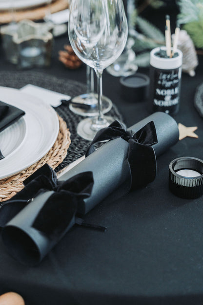 Decorative table setting with black napkins, glasses, and black candles on a black tablecloth with black Christmas crackers and black velvet bows by Noble & Nordic.