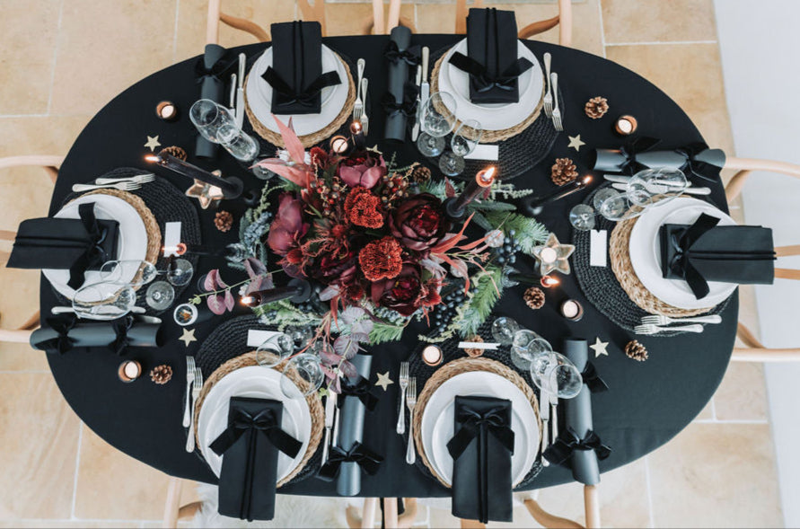 Elegant black oval table set with white plates, black napkins, and decorative Christmas centrepiece by Noble & Nordic.