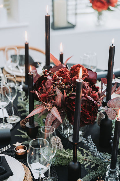 Decorative table setting with black candles, burgundy Christmas flower bouquet, and fir pine cone Christmas garland on a black tablecloth by Noble & Nordic.