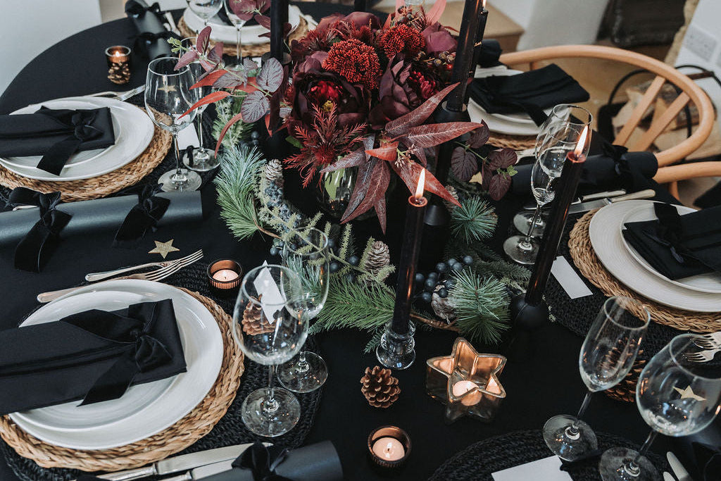 Elegant table setting with black tablecloth, white plates, black napkins, and burgundy floral bouquet decorative centrepiece, black crackers with black velvet bow.