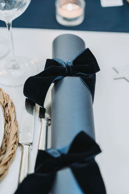 Navy Christmas cracker with navy velvet bow tie on a Christmas table setting with cutlery and glasses.