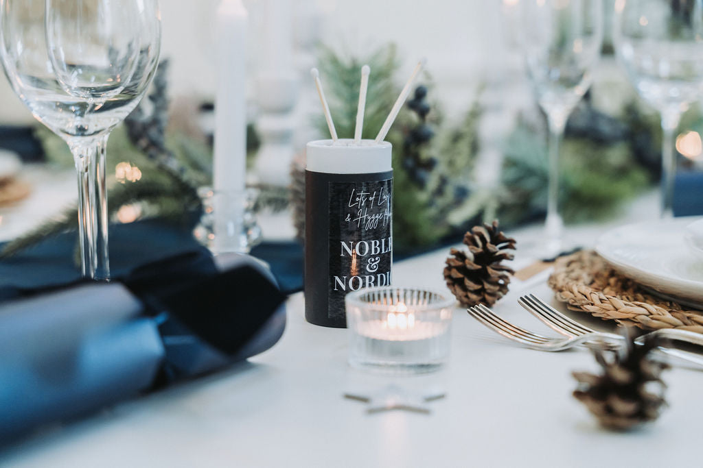 Nordic-navy themed table setting with white candles, pinecones garland, and a branded matchstick box, designed by Noble & Nordic.