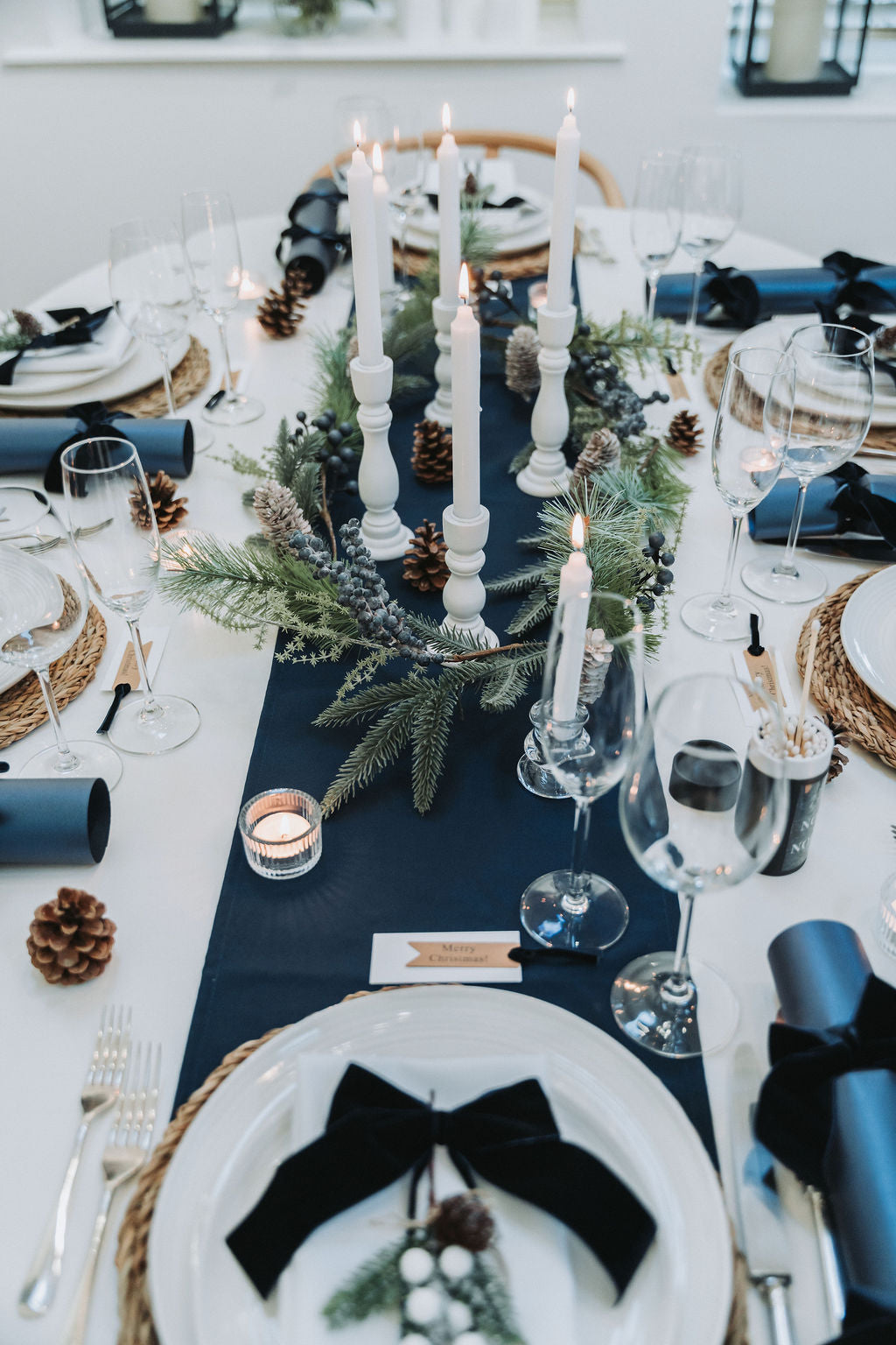 Elegant navy Christmas table setting with white candles, Christmas fir pine garland, and navy blue Christmas crackers with navy velvet bows, designed by Noble & Nordic.