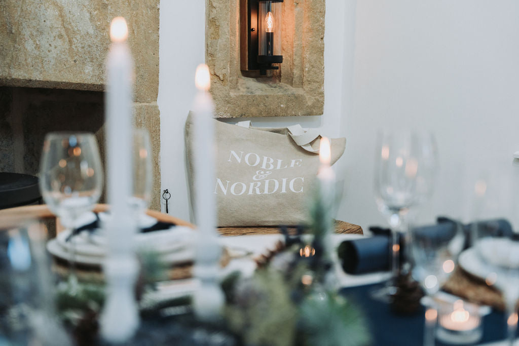 Dining table setting with white candles and a 'Noble & Nordic' branded jute bag in the background, designed by Noble & Nordic.