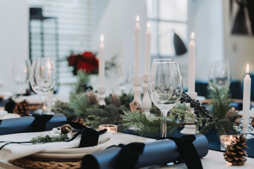 Decorative navy Christmas table setting with white candles, glasses, navy Christmas crackers with navy velvet bow and Christmas fir pine garland in a blurred indoor setting, designed by Noble & Nordic.