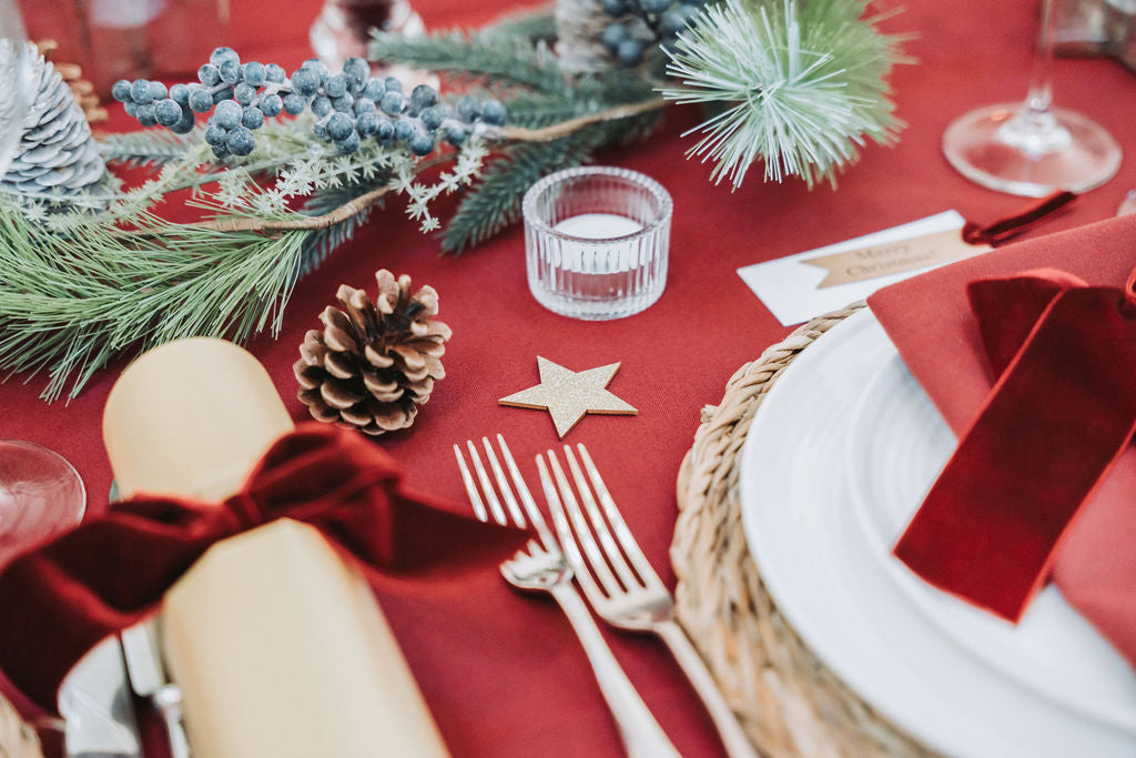 Burgundy Decorative table setting with darl red tablecloth, pine cone fir garland, and Christmas decorations, designed vy Noble & Nordic.