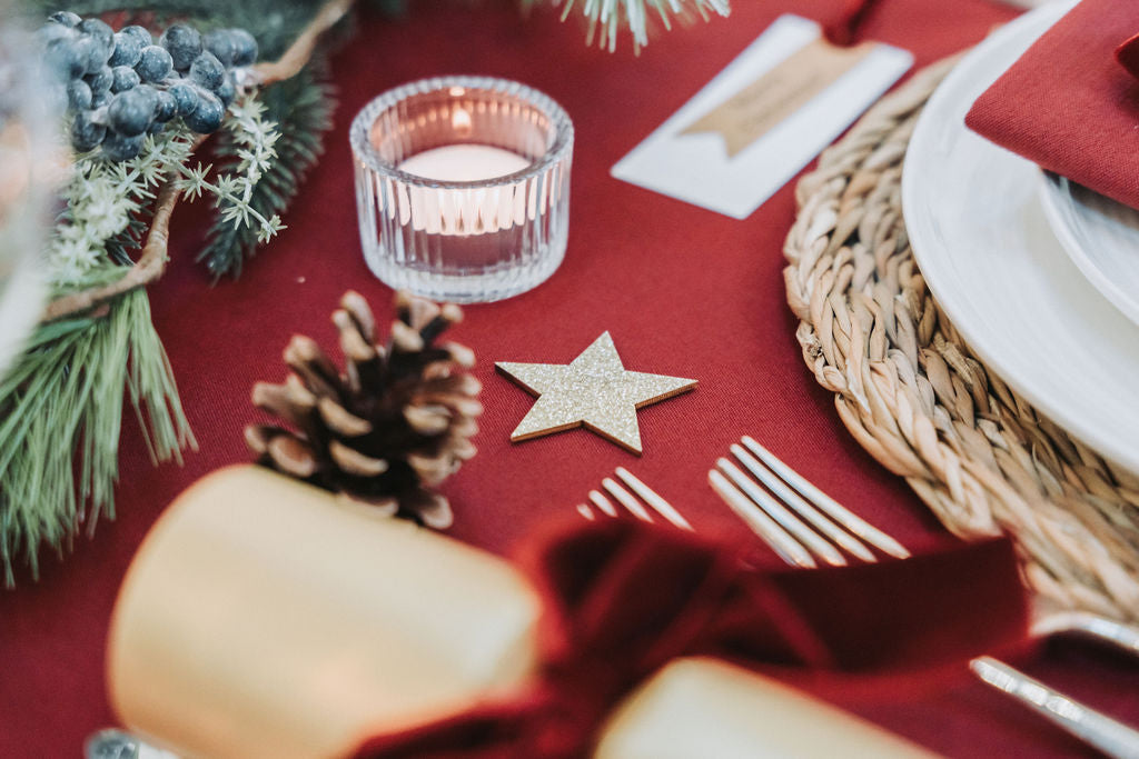 Christmas-themed table setting with dark red tablecloth, Christmas fir pine cones garland, wooden star decorations, and dark red candles, designed by Noble & Nordic.
