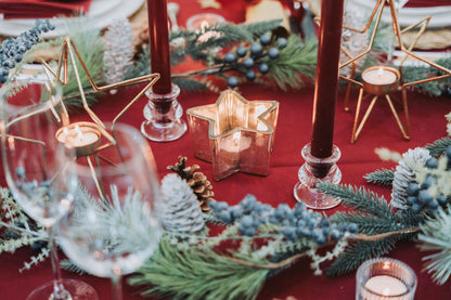 Burgundy Christmas decorative table setting with red candles, glasses, and fir pine cones garland on a dark red tablecloth, by Noble & Nordic.