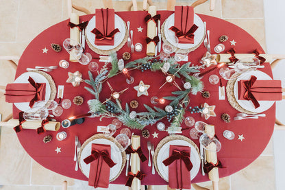 Burgundy decorative Christmas table setting with dark red tablecloth, dark red napkins, and centerpieces with Christmas fir pine cone garland, and gold Christmas crackers with dark red velvet bows, designed by Noble & Nordic.