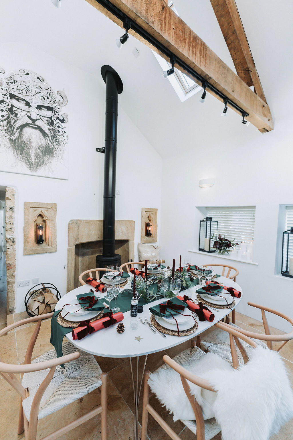 View of festive table setting with forest green table linens, pine foliage Christmas garland, red candles, pearlised red crackers with burgundy velvet bows by Noble & Nordic.