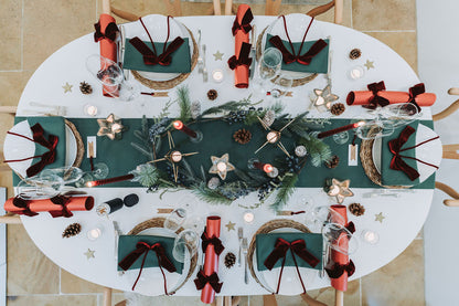 Overhead view of festive forest green table setting with green table linens, pine cone fir Christmas garland and starry tealight candleholder, red pearlised crackers with burgundy velvet bow, by Noble & Nordic.