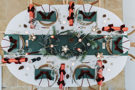Overhead view of festive forest green table setting with green table linens, pine cone fir Christmas garland and starry tealight candleholder, red pearlised crackers with burgundy velvet bow, by Noble & Nordic.