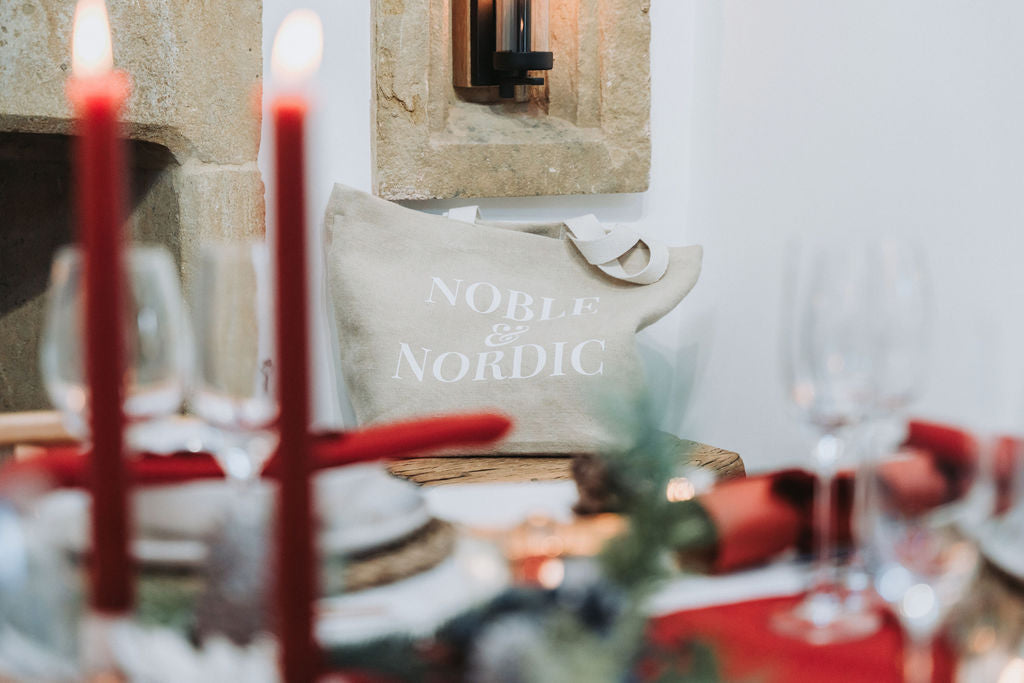Decorative setting with candles, a pillow, and glasses on a table with Noble & Nordic jute bag.