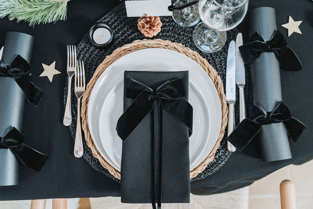 Nordic Noir Christmas table setting with black velvet ribbon gift box, black crackers, rattan placemat and pinecone detail — sophisticated festive styling by Noble & Nordi