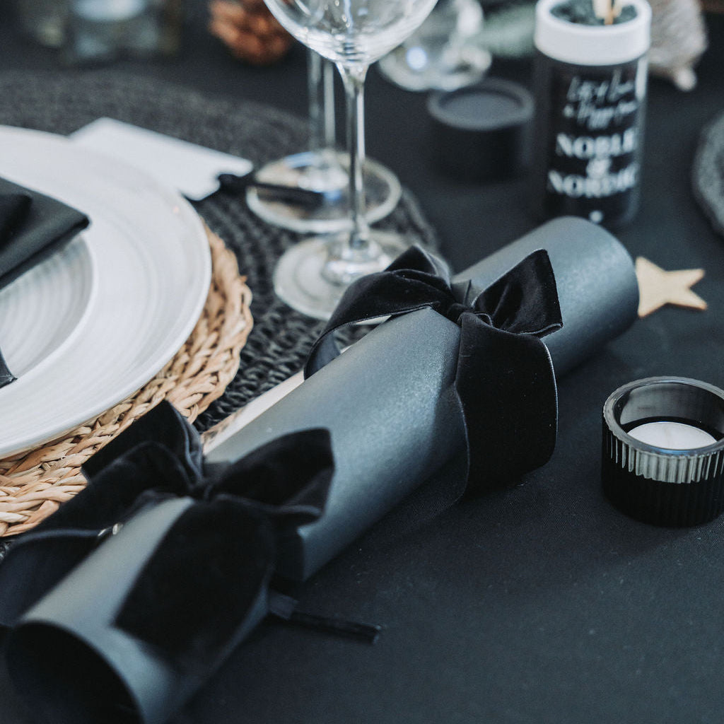 Black Christmas tablescape with black velvet Christmas crackers, wrapped gift box, pinecones and glassware — elegant Nordic festive styling by Noble & Nordic.