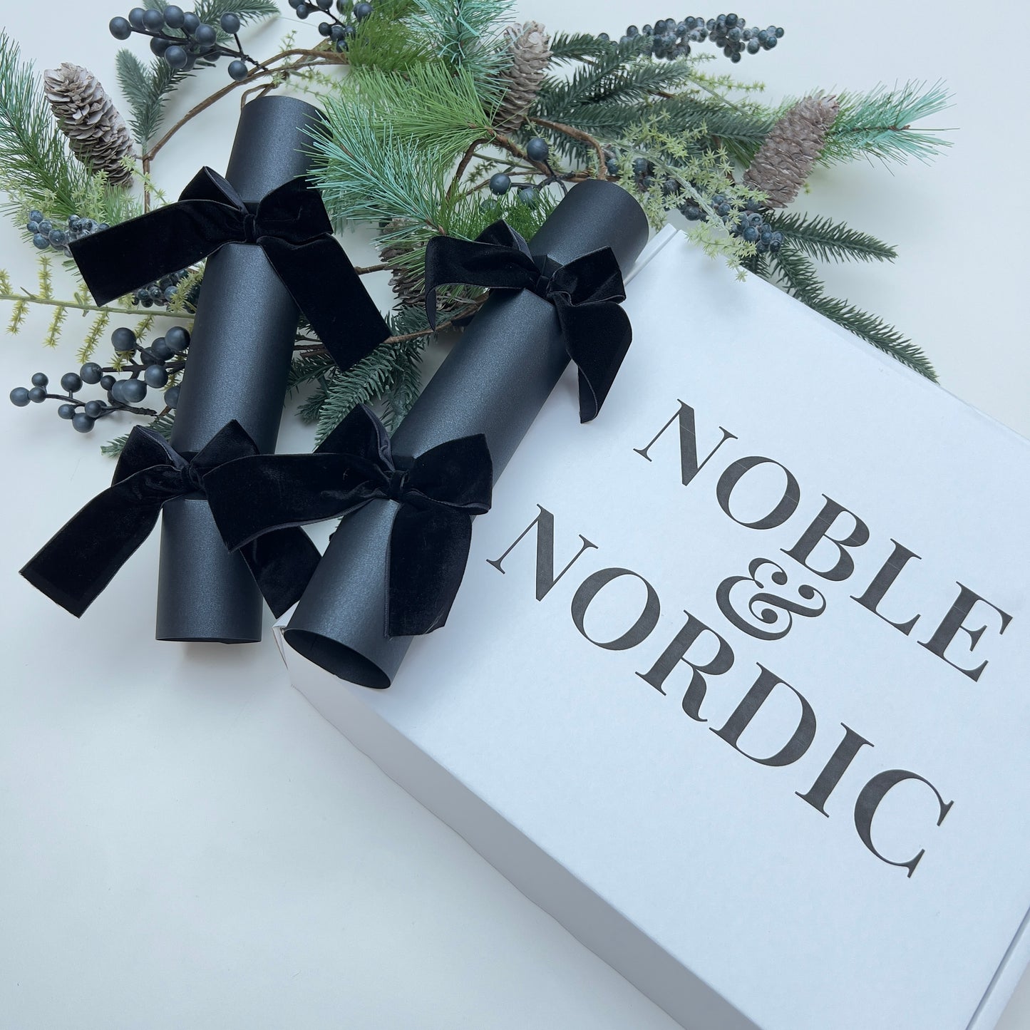 Gift box with black Christmas cracker with black velvet bows and 'Noble & Nordic' branding, surrounded by Christmas fir pine cone garland.
