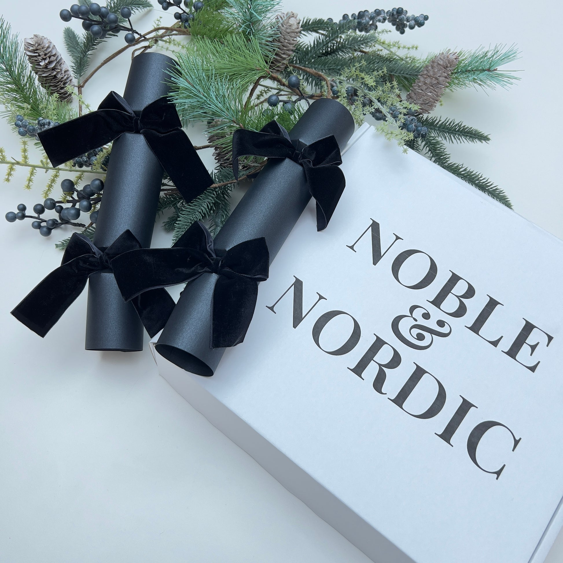 Gift box with black Christmas cracker with black velvet bows and 'Noble & Nordic' branding, surrounded by Christmas fir pine cone garland.