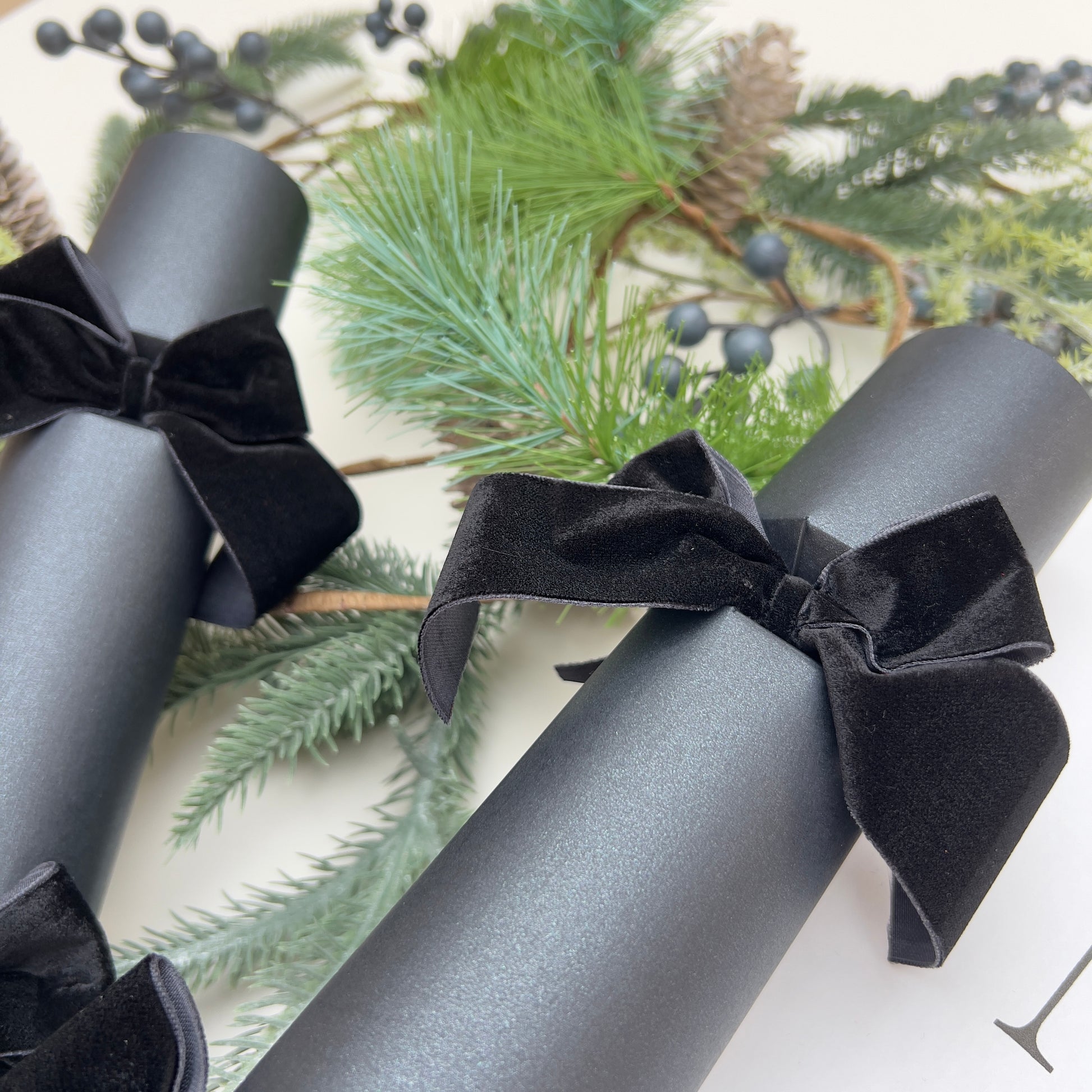 Black Christmas crackers with black velvet bows and Christmas fir pine cone garland in the background by Noble & Nordic.
