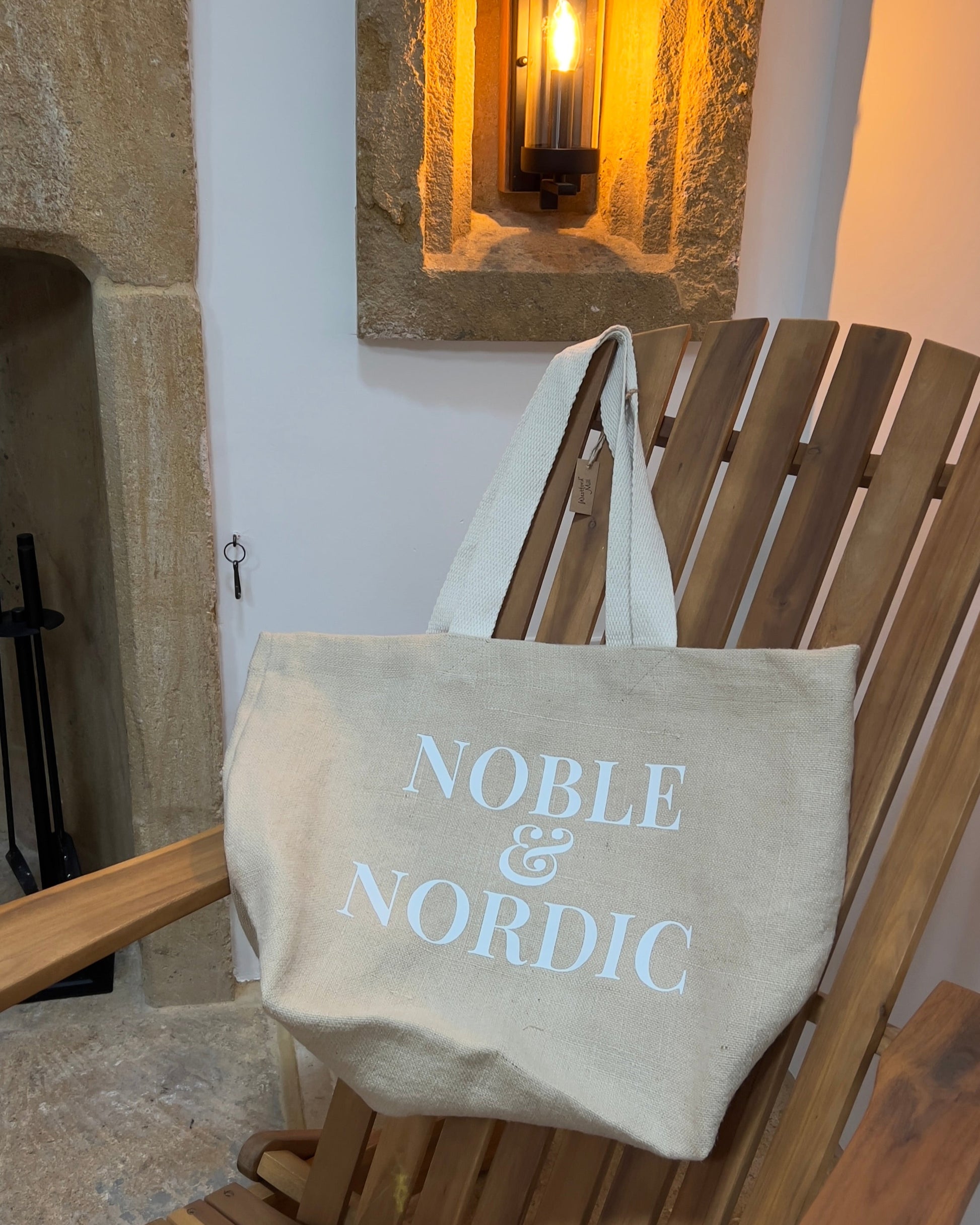 Beige tote bag with 'Noble & Nordic' text on a wooden chair against a stone wall background designed by Noble & Nordic.