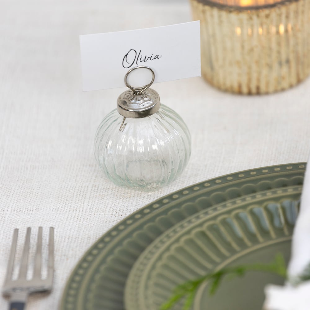 Ribbed glass place card holder with silver metal top and handwritten name tag reading “Olivia,” styled on an elegant neutral table with green dinnerware and a softly glowing candle.