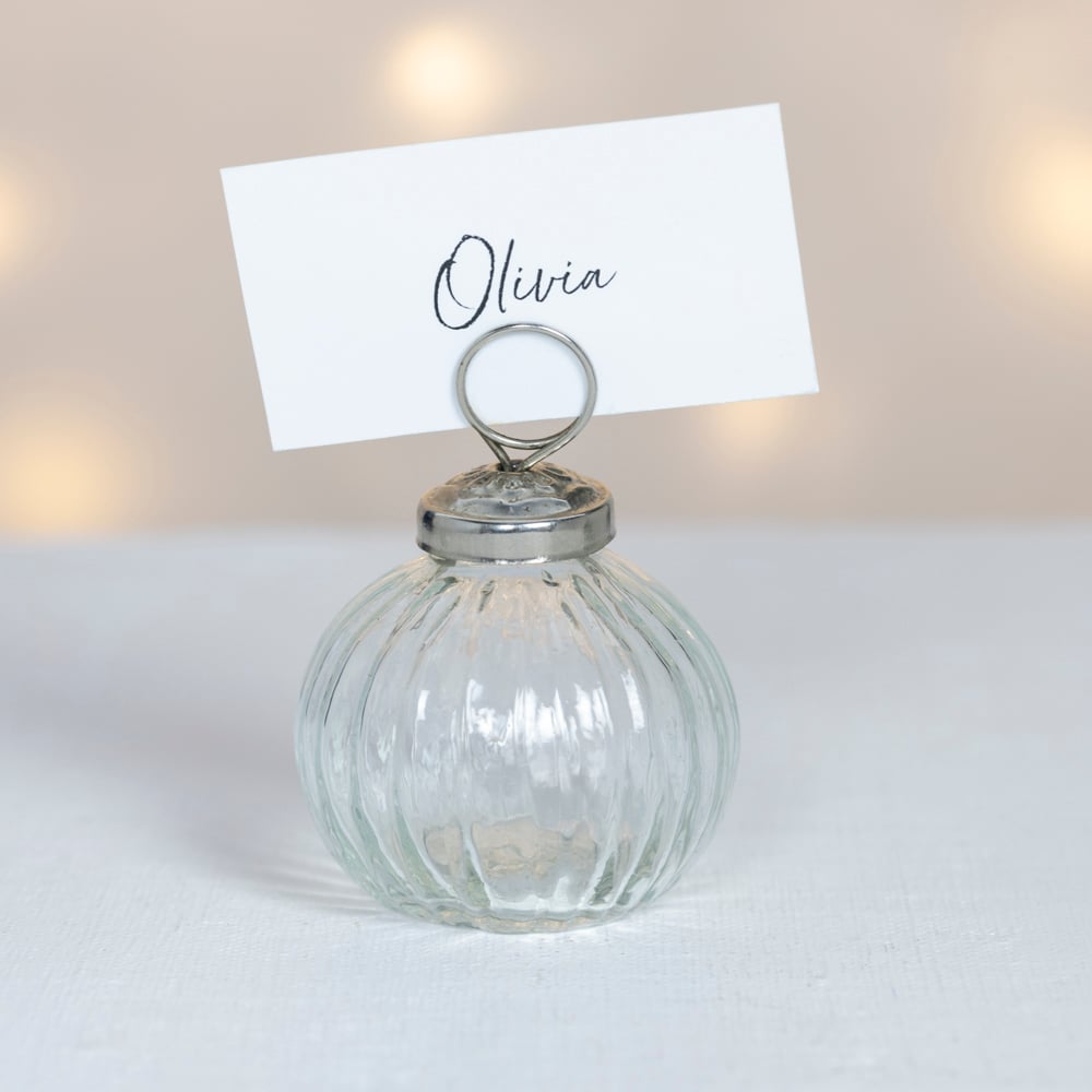 Clear ribbed glass place card holder with silver top and handwritten name card, photographed against a soft neutral background with warm glowing lights.